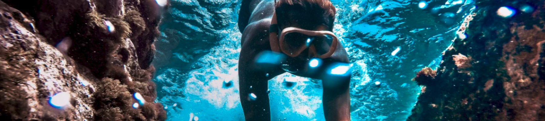 A person snorkeling underwater, surrounded by rocky formations and vibrant blue water, wearing goggles in a sunlit aquatic scene.