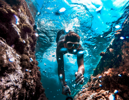 A person snorkeling underwater, surrounded by rocky formations and vibrant blue water, wearing goggles in a sunlit aquatic scene.