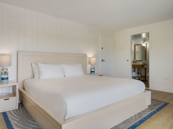A modern bedroom features a white bed, two side tables with lamps, and a bathroom visible through an open door.