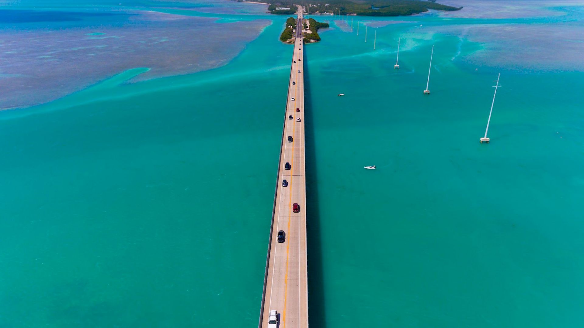 A long pier extends over turquoise waters to a distant land, with a few cars driving along, and wind turbines dotting the scene.
