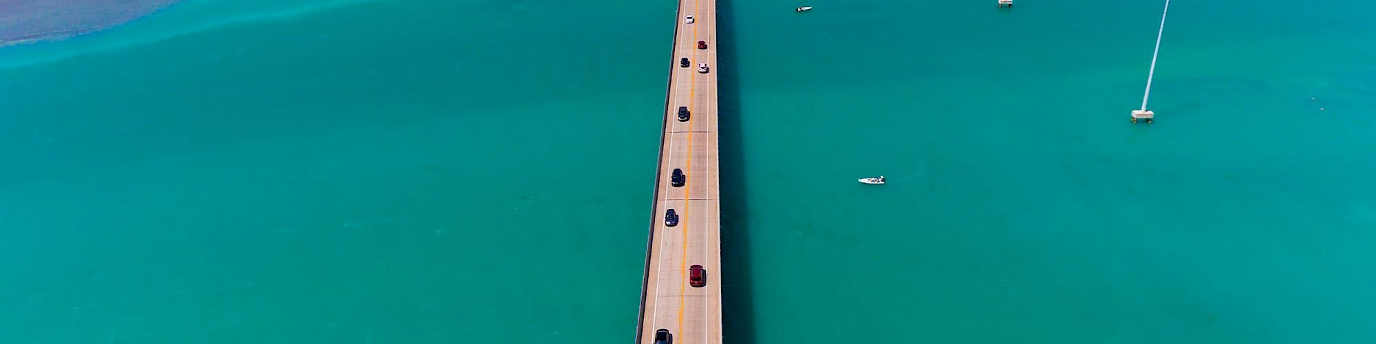 A long pier extends over turquoise waters to a distant land, with a few cars driving along, and wind turbines dotting the scene.