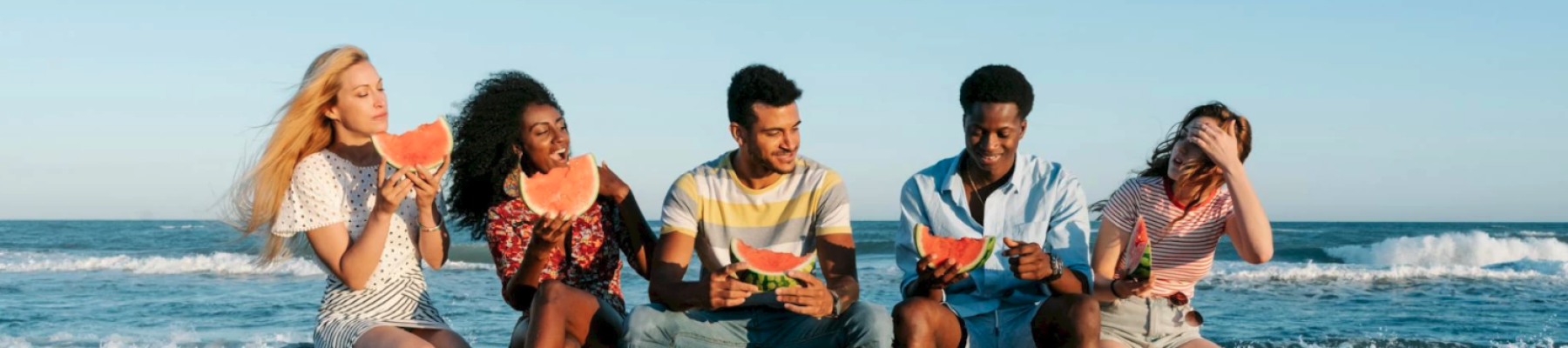 Five friends sit on a beach eating watermelon together as waves roll in, enjoying sunny weather and each person smiling or laughing.
