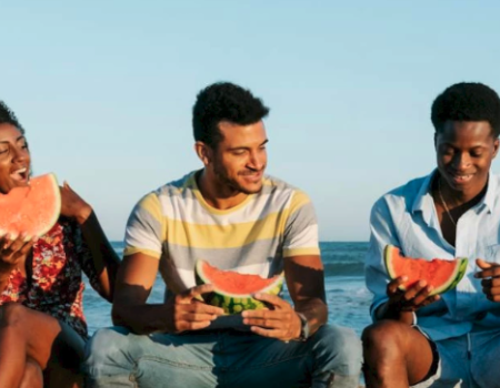 Five friends sit on a beach eating watermelon together as waves roll in, enjoying sunny weather and each person smiling or laughing.