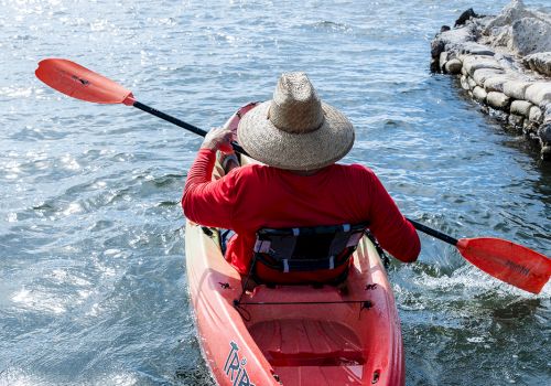 A person wearing a hat paddles a red kayak toward the open sea with calm blue water and a rocky jetty in the distance.