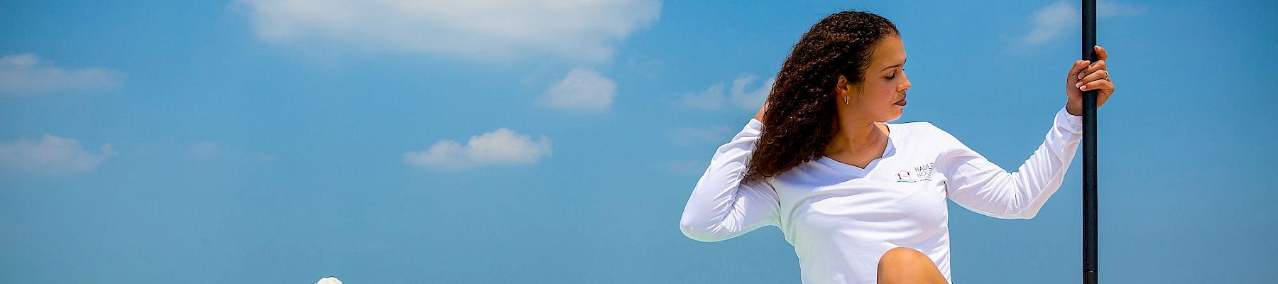 Woman in white top sits on a paddleboard at the beach, holding a paddle, with blue sky and water in the background.