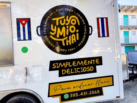 Cuban-style food truck with a black circular logo, yellow &ldquo;Simplemente Delicioso&rdquo; sign, Cuban flags, and contact info on the back of a white trailer.