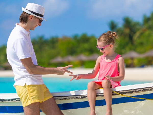 A man and a young girl share a high-five on a sunny beach or boat, by the water, wearing summer clothes and sunglasses.