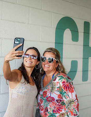 Two friends take a selfie in front of a wall that says &ldquo;CHILL&rdquo; wearing sunglasses, smiling and posing together.