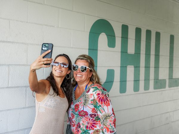 Two friends taking a selfie against a wall with a large &ldquo;CHILL&rdquo; mural, both wearing sunglasses and smiling as they pose, ending with a period.