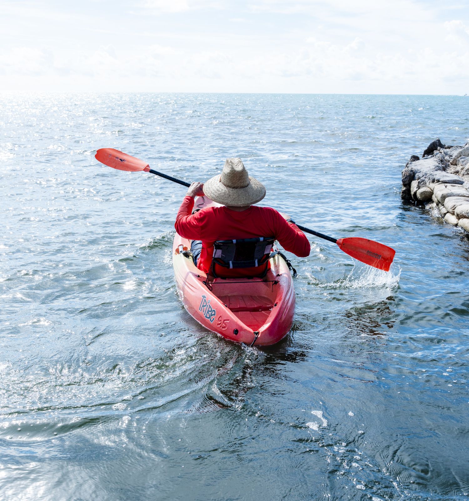 A person in a red kayak paddles away from shore into calm blue water, wearing a hat, near rocky breakwater, bright sky above.