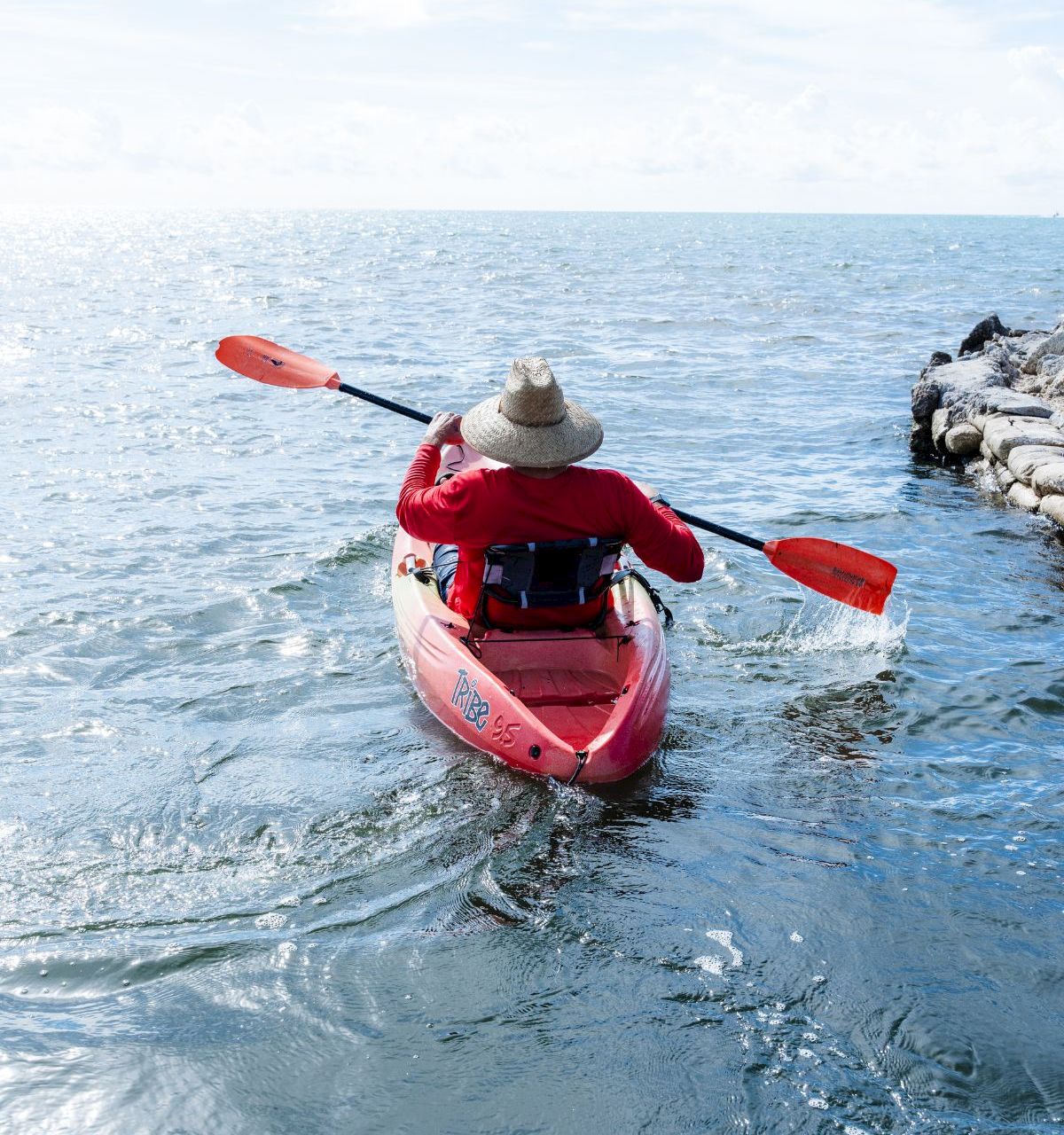 A kayaker in a red boat paddles on open water beside rocks under a bright, cloudy sky.