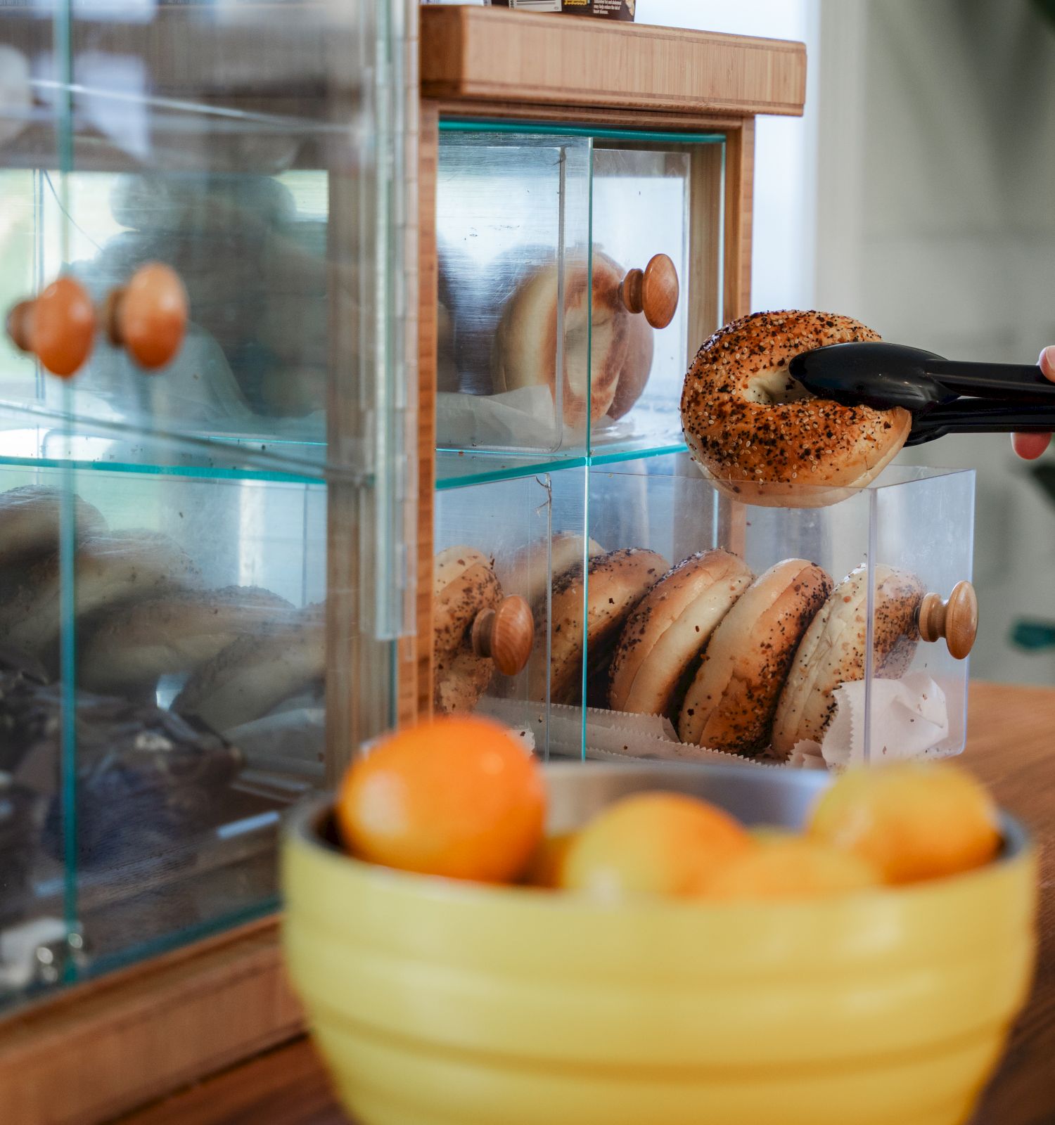 A bakery display with glass cases, pretzels and bagels inside, and a person grabbing a bagel with tongs near a bowl of oranges.