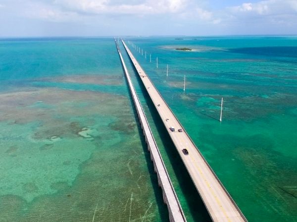 A long bridge stretches over turquoise sea, connecting land to distant horizon, with clear shallow waters and light poles lining its sides.