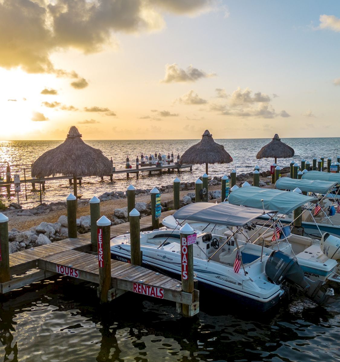 A sunset marina with white boats on a dock, thatched palapas, and rental/tour signage by the calm ocean.