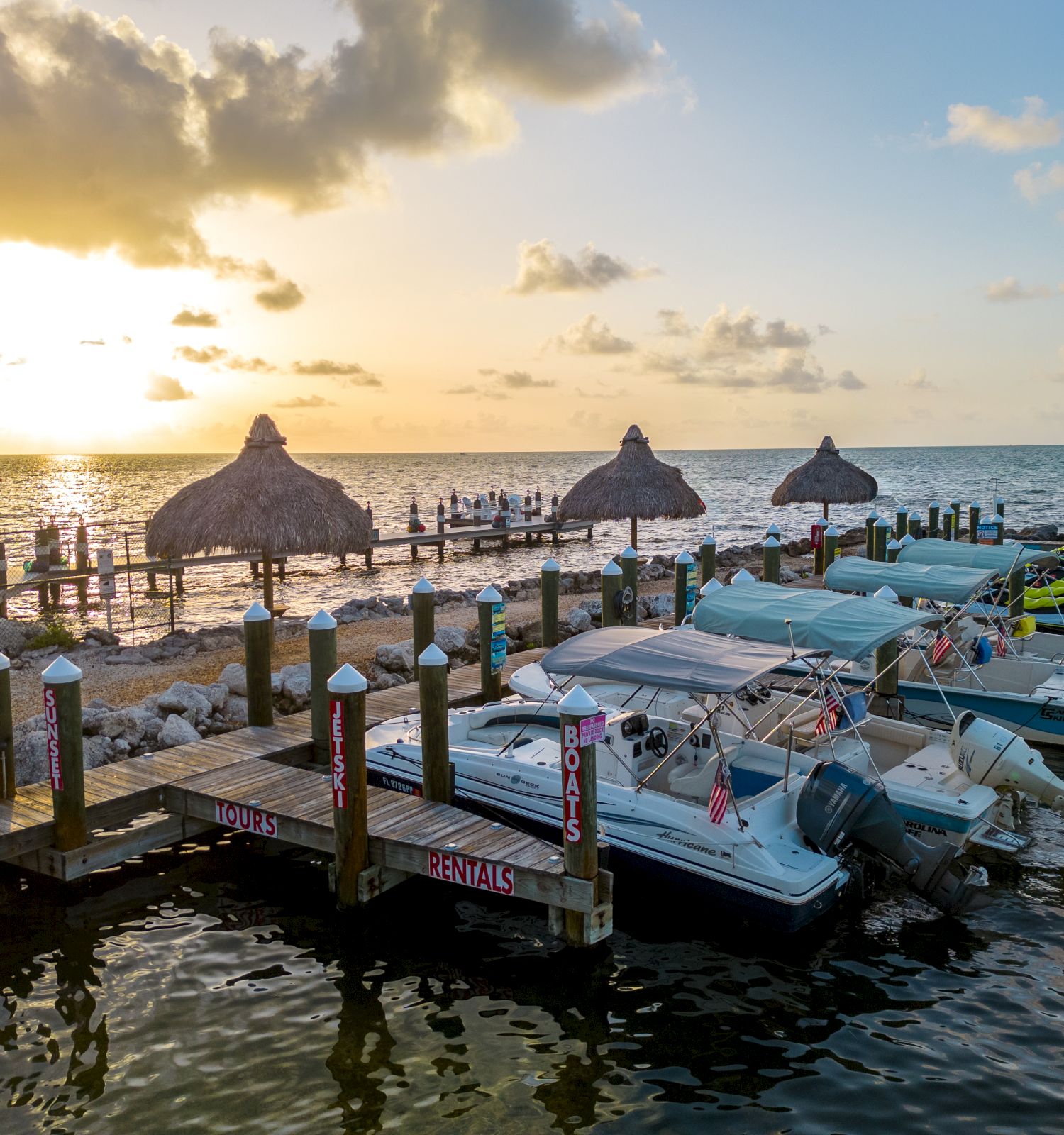 A tropical pier at sunset: calm sea, thatched shade huts, and boats lined up, ready for sunset rides and island vibes.