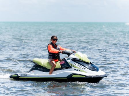 A person riding a green and white jet ski on calm ocean water, wearing a life vest and sunglasses, near shore.