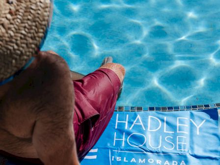 A person wearing a straw hat sits by a bright blue pool, with a blue Hadley House towel or sign on the edge. All ending with a period.