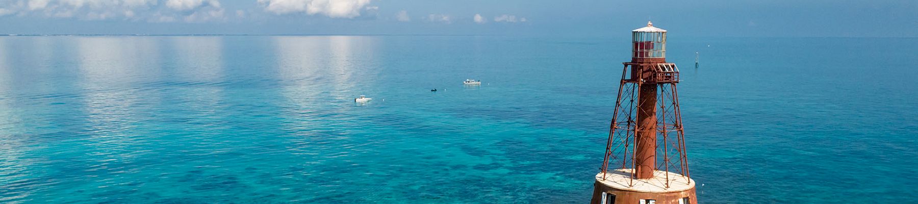 A red-and-white lighthouse tower stands in clear turquoise sea, supported by metal stilts, with a distant horizon and a few puffy clouds above.