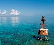 A red-and-white lighthouse tower stands in clear turquoise sea, supported by metal stilts, with a distant horizon and a few puffy clouds above.