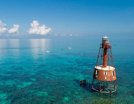 A red-and-white lighthouse tower stands in clear turquoise sea, supported by metal stilts, with a distant horizon and a few puffy clouds above.