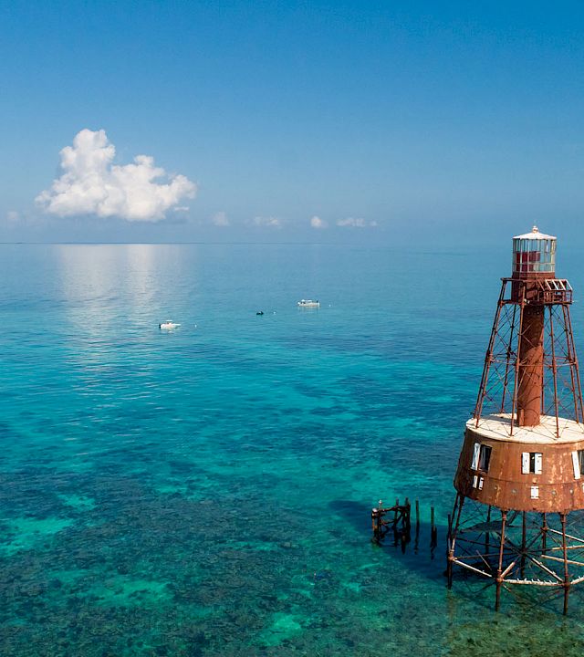 A red-and-white lighthouse tower stands in clear turquoise sea, supported by metal stilts, with a distant horizon and a few puffy clouds above.
