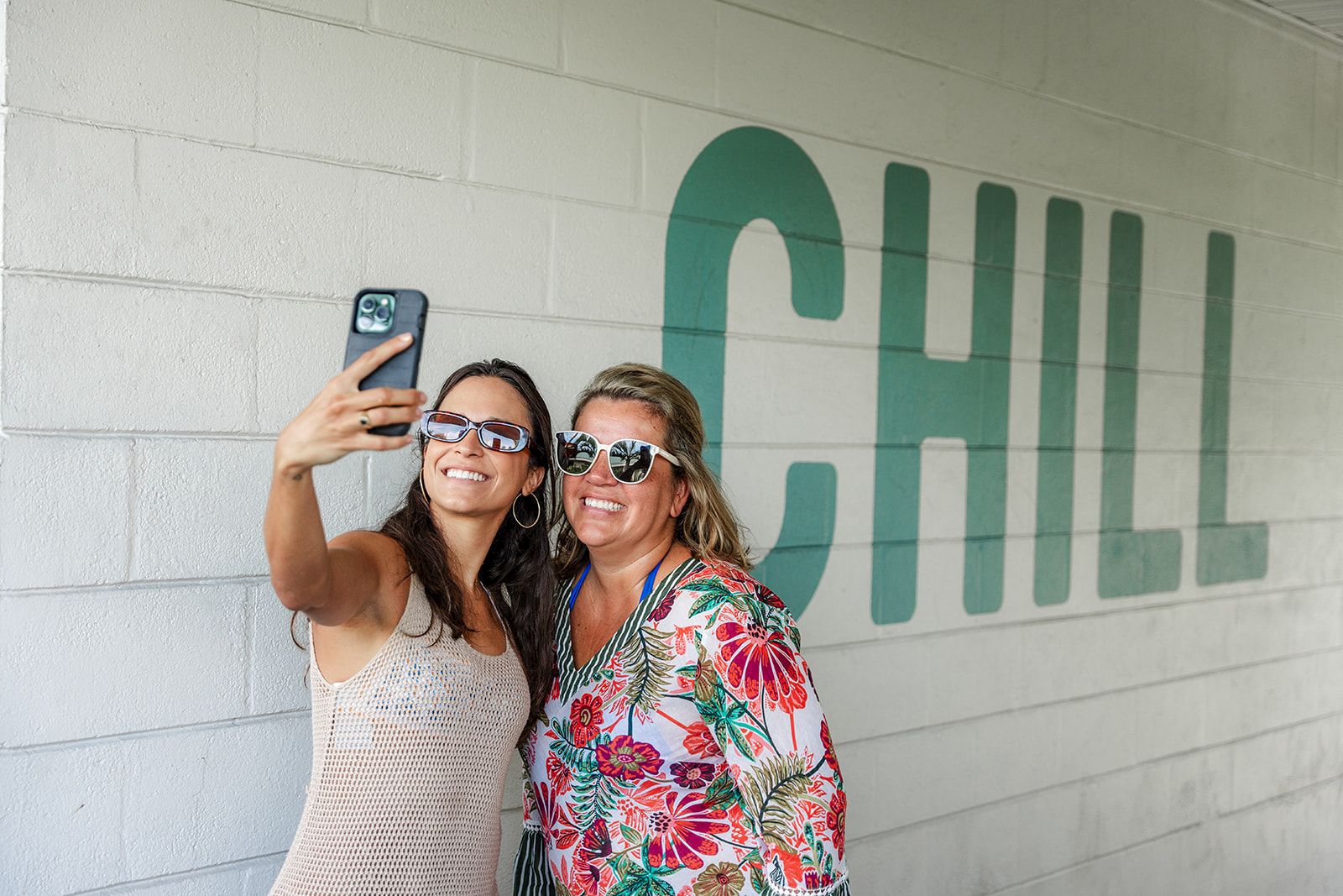 Two women in sunglasses pose for a selfie, smiling and holding up phones in front of a green wall mural.