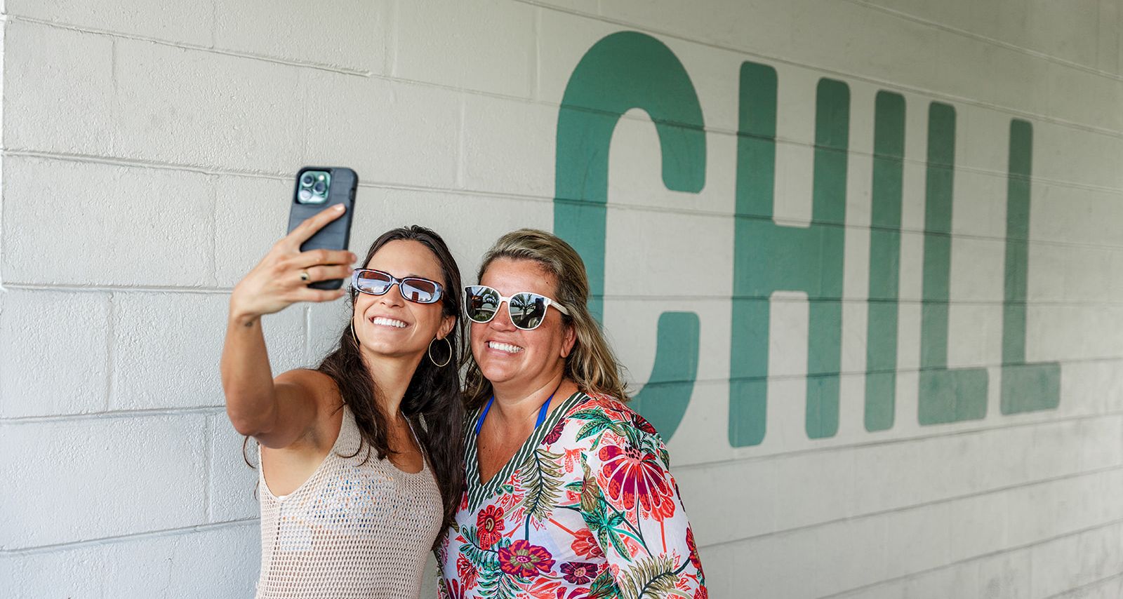 Two women in sunglasses pose for a selfie, smiling and holding up phones in front of a green wall mural.