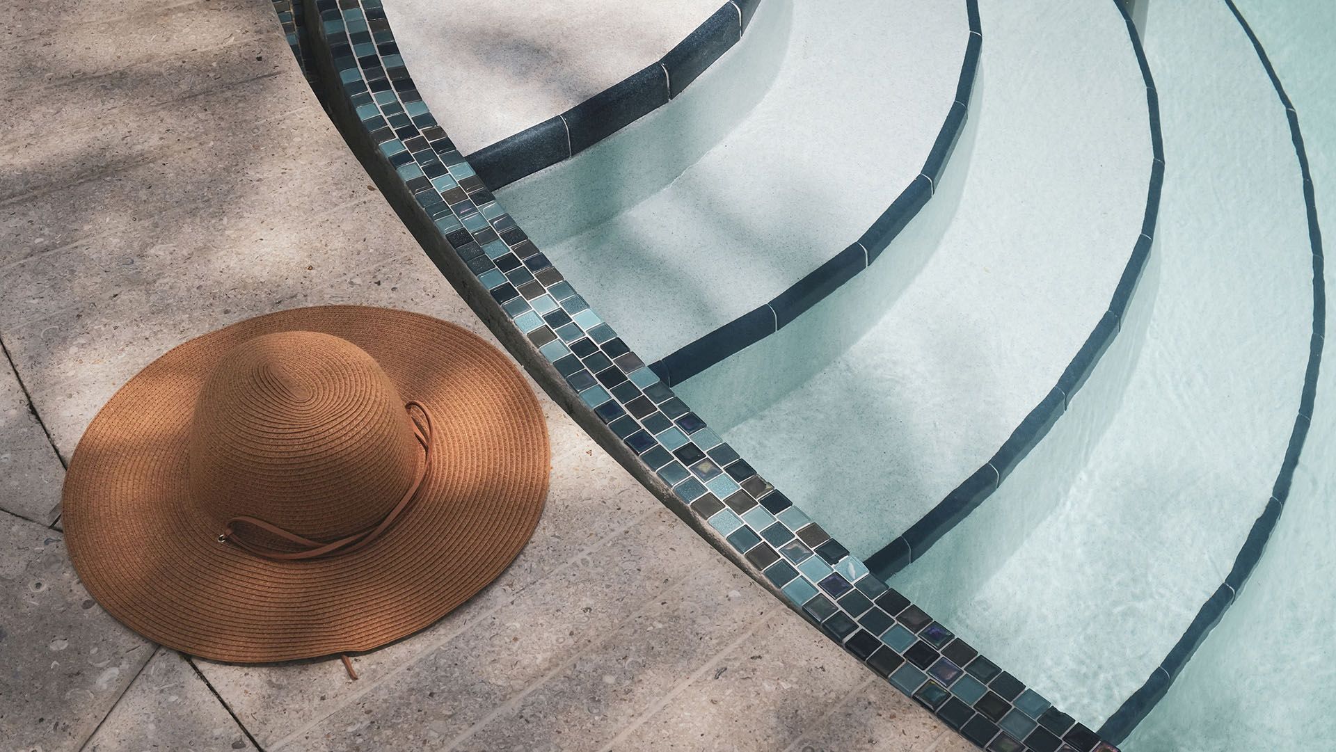 A brown wide-brimmed hat rests by a curved pool edge lined with blue mosaic tiles and light aqua water.