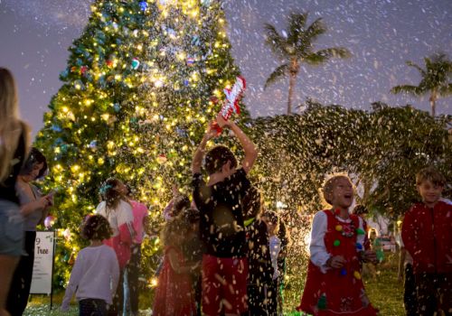 Kids in festive outfits run past a glowing Christmas tree as confetti falls in a snowy night celebration.