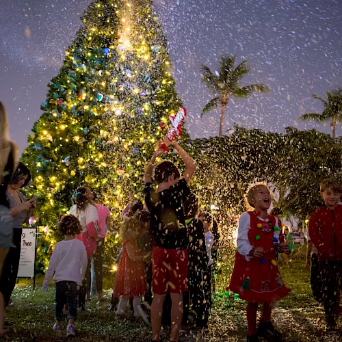 Kids in festive outfits run past a glowing Christmas tree as confetti falls in a snowy night celebration.