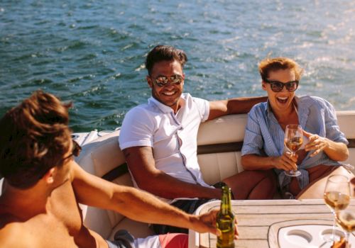 Friends on a sunny boat laugh and toast with drinks while chatting by the waves in sunglasses.