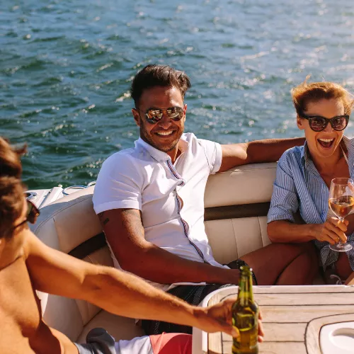 Friends laugh on a sunny boat by the sea, clinking champagne glasses and enjoying drinks together at sunset.