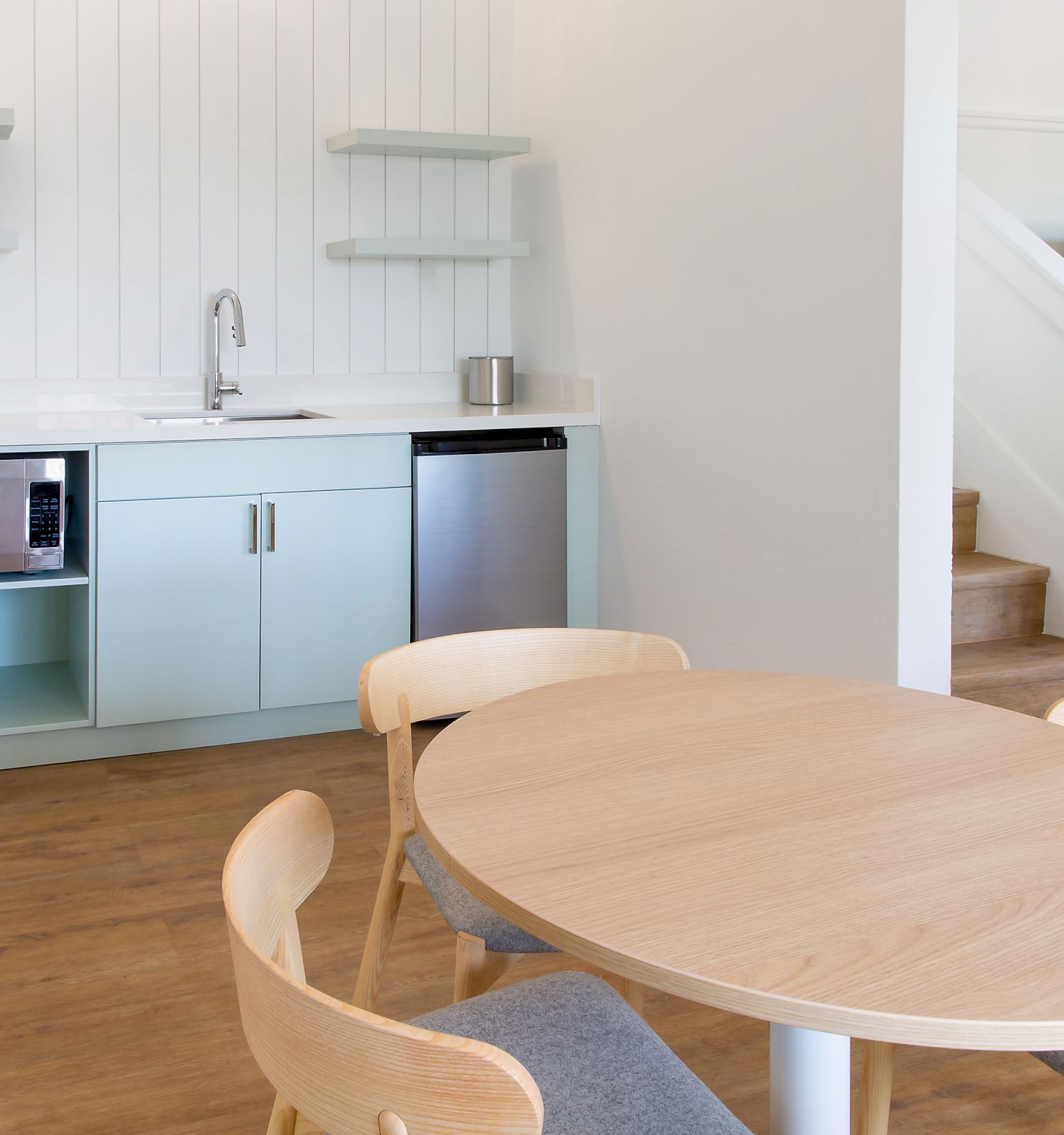 The image shows a minimalist kitchen and dining area with a round table and chairs, a kitchenette with appliances, and stairs leading up.