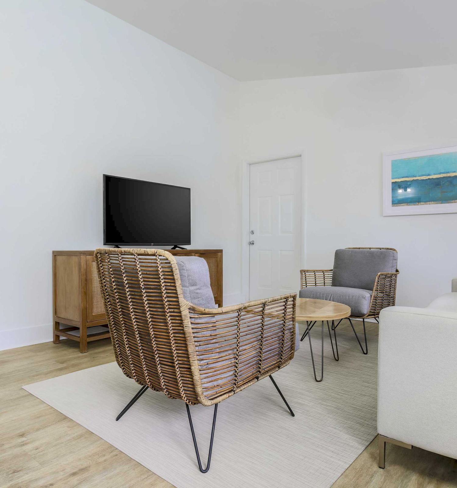 A modern living room with a TV, two wicker chairs, a sofa, a round coffee table, a lamp, and a coastal-themed wall art. A staircase is on the left.