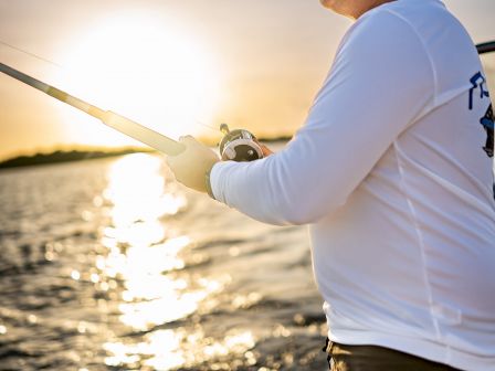 A person is fishing on a boat during sunset, wearing sunglasses and a long-sleeve shirt. The sun reflects on the water, creating a serene scene.