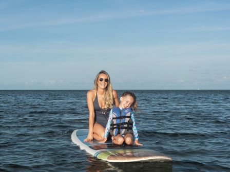 A woman and child are sitting on a paddleboard in calm ocean waters under a clear sky, both wearing swimsuits and the child wearing a life jacket.