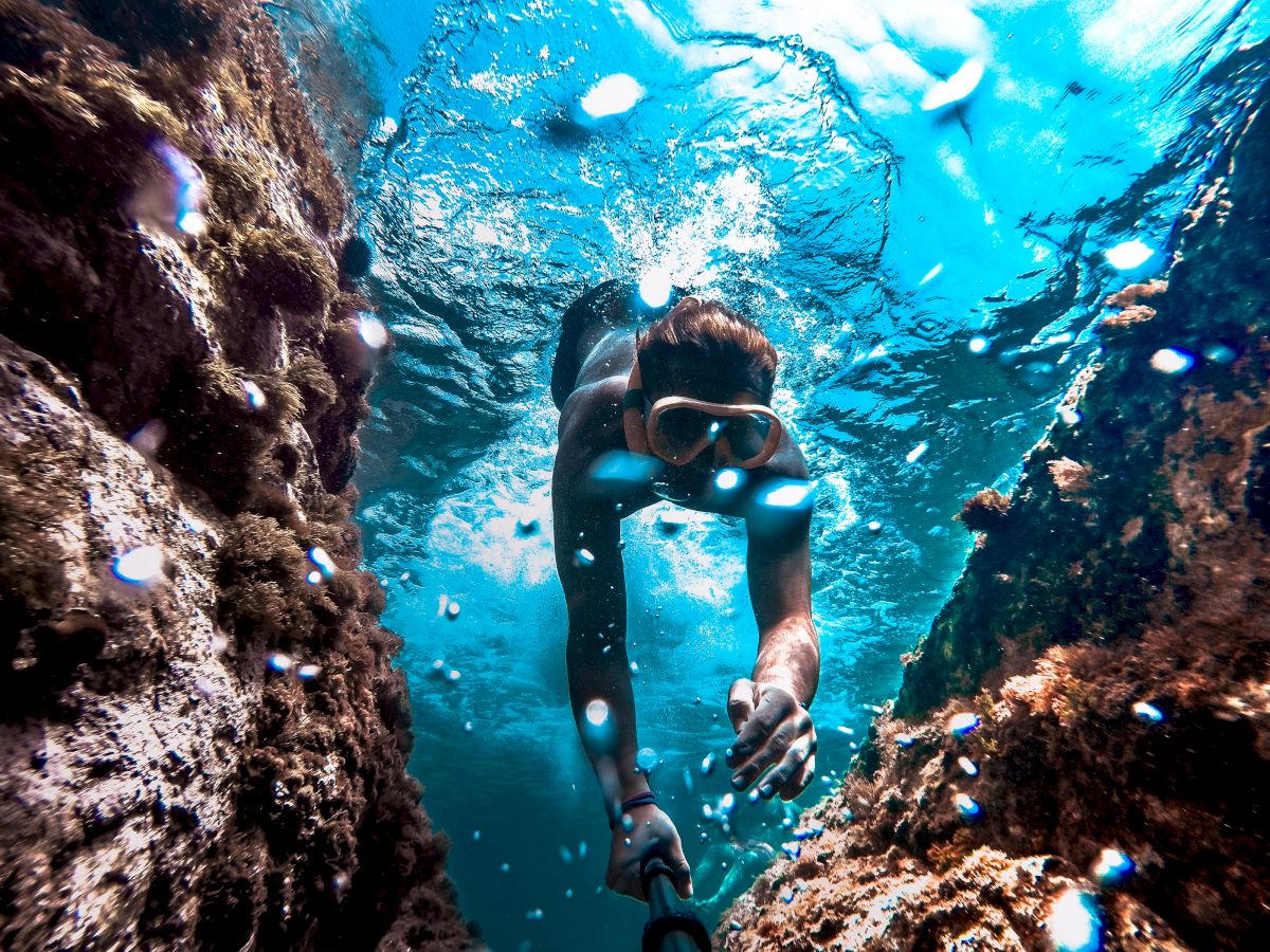 A person wearing a mask is snorkeling underwater between rocky formations, with sunlight filtering through the clear blue water.