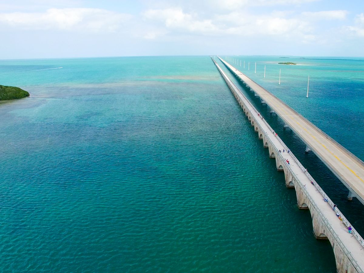 A long bridge stretches over turquoise waters leading to small islands under a partly cloudy sky, featuring lush greenery and utility poles.