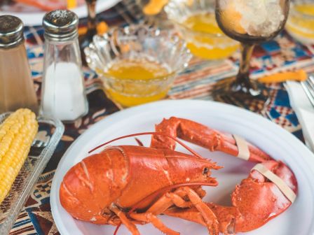 A plate with a cooked lobster, corn, and drinks are on a colorful tablecloth, surrounded by condiments and more food items.