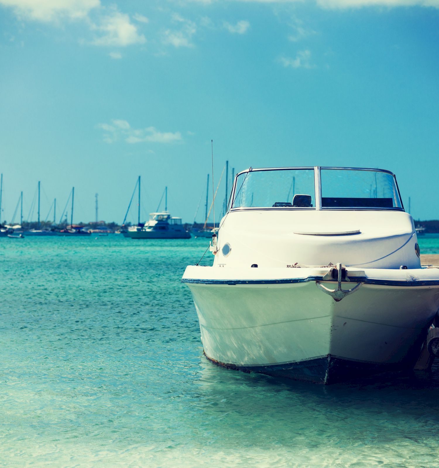 A white motorboat is docked beside a pier, with numerous sailboats anchored in the turquoise water in the background.