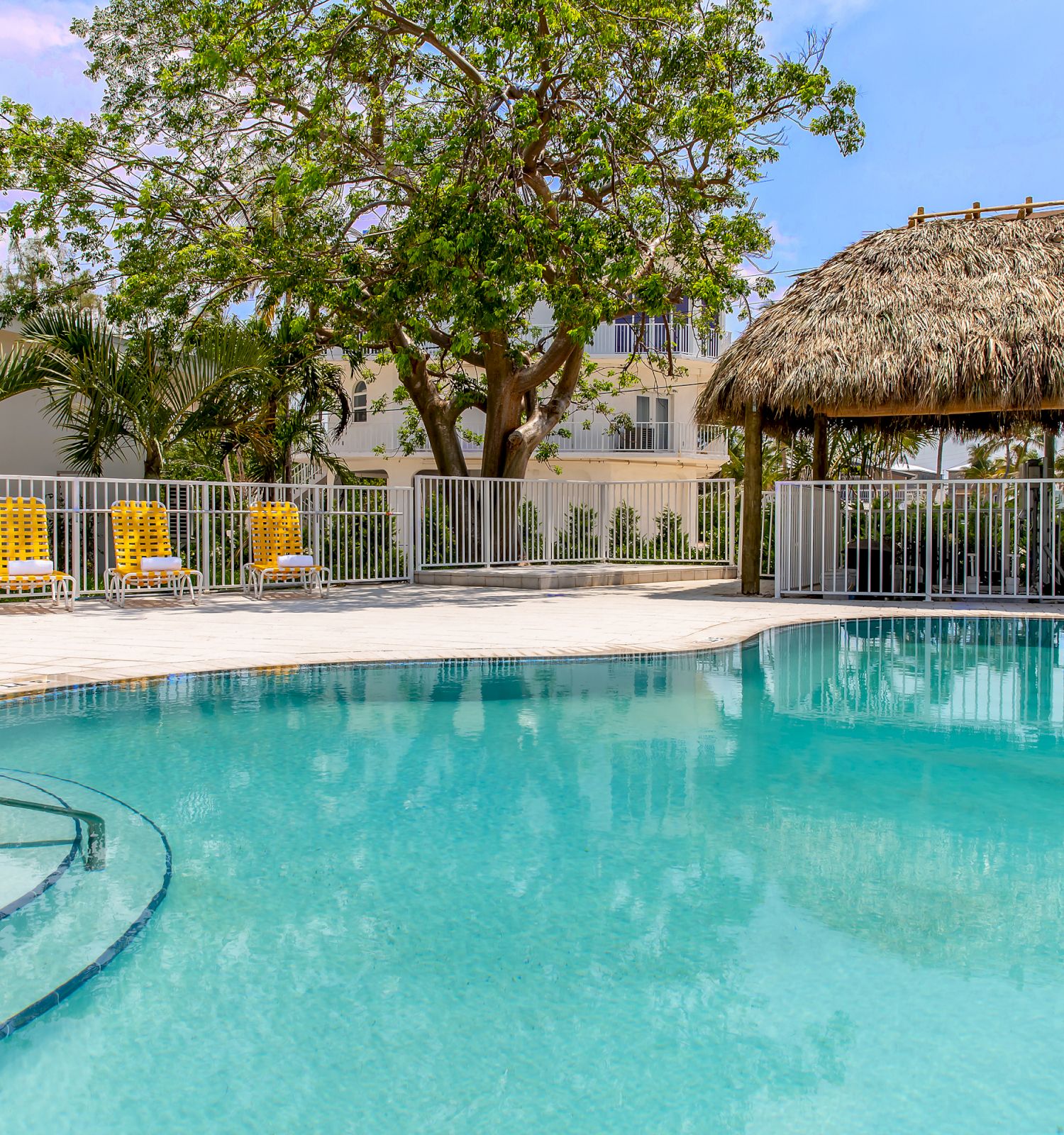 A swimming pool with steps and railings, a thatched-roof cabana, yellow chairs, and a fence, surrounded by palm trees and a building in the background.