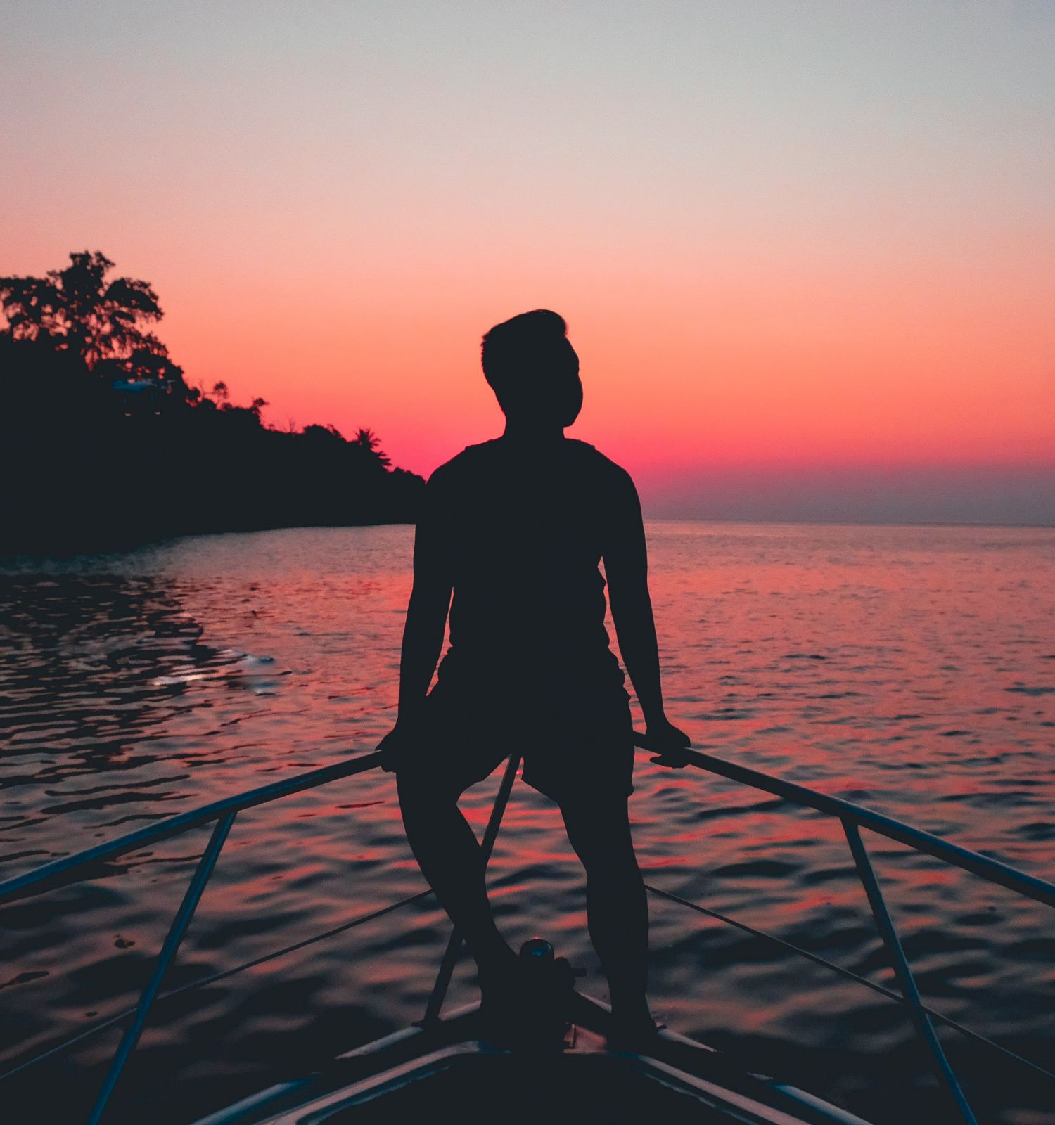 A silhouette of a person standing on a boat's bow, overlooking a calm sea at sunset with trees on the left and vibrant colors in the sky.