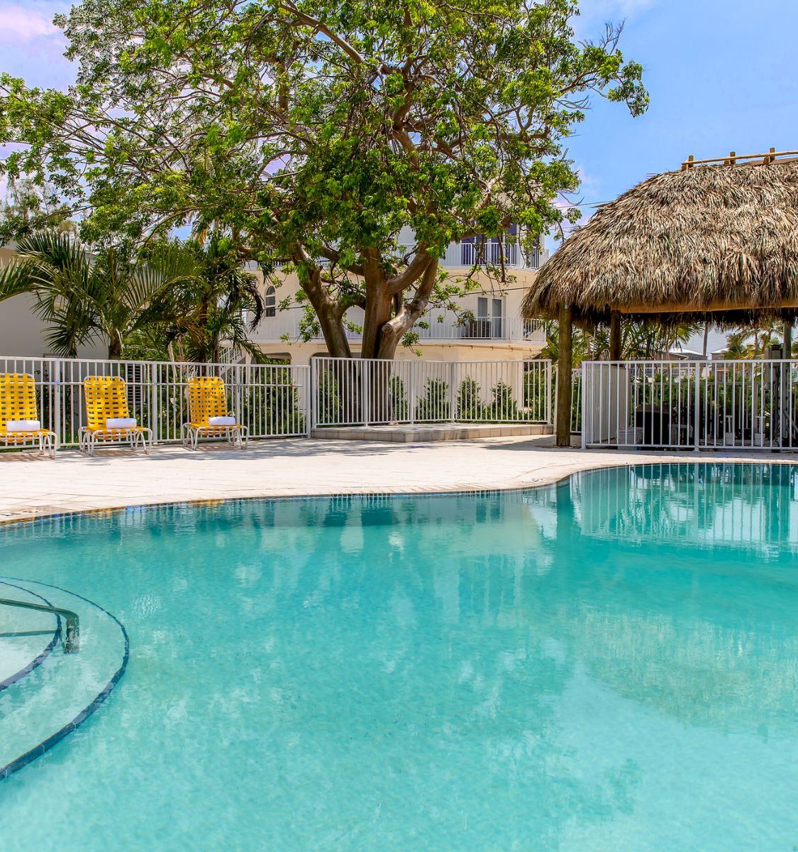 Sunny resort pool with turquoise water, lounge chairs, and a thatched palapa by tropical trees and a white building.