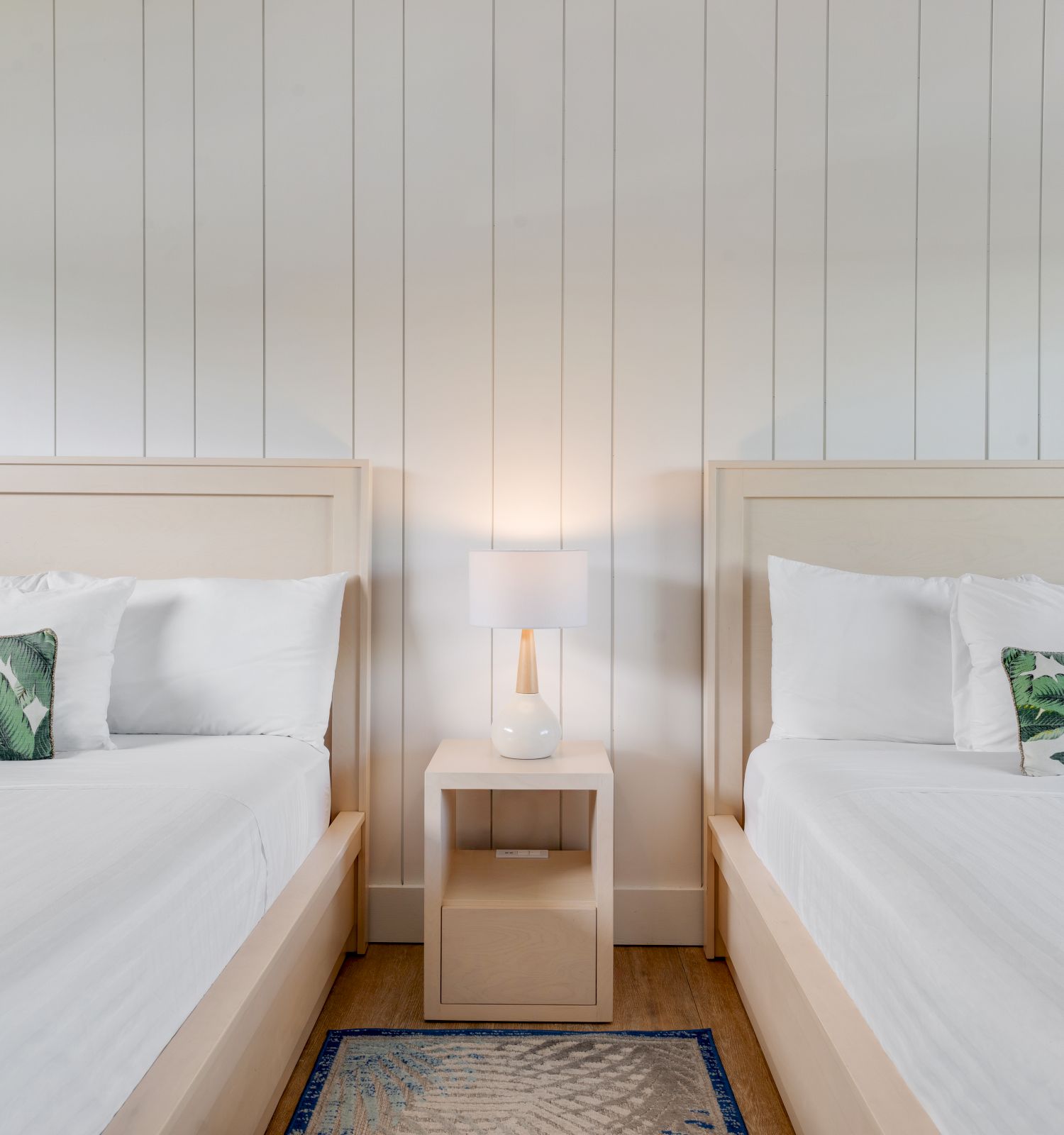 This image shows a room with two neatly made beds, each with white bedding and a green leaf-patterned pillow, separated by a nightstand with a lamp.